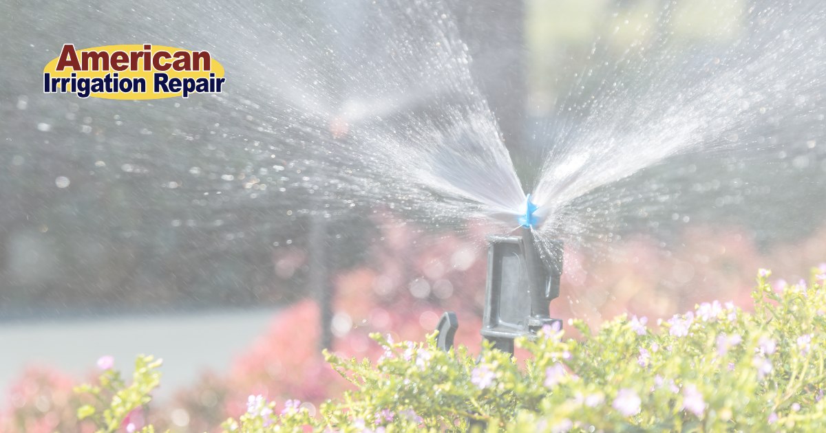 A Rain Bird sprinkler system in a yard, watering the grass. The Rain Bird sprinkler head, mounted on a tall pipe, sprays a fine mist of water over the green grass, ensuring even coverage. Surrounding the grass are plants and flowers, adding color to the garden.