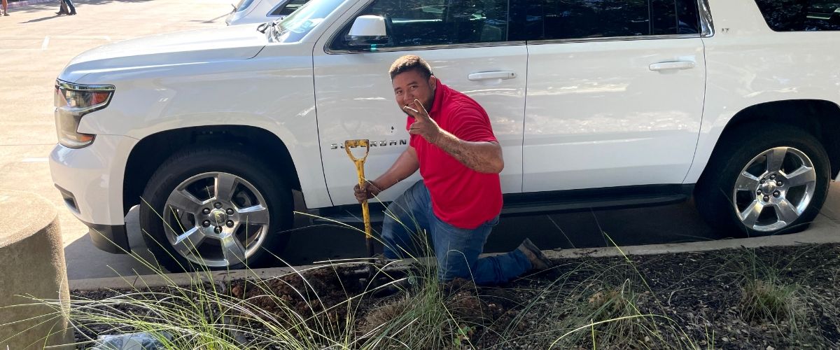 A sprinkler technician in action, rebuilding a sprinkler system in a parking lot. The technician is inspecting and replacing pipes, valves, and sprinkler heads, with a car visible in the background