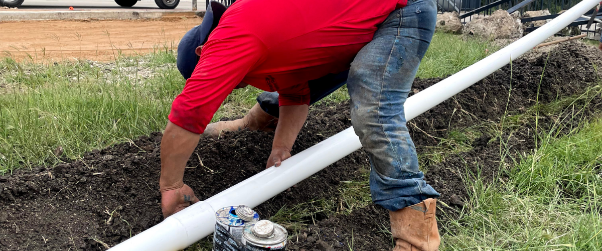 A person wearing a red shirt is positioned over an irrigation pipe as they work on installing a lawn sprinkler system