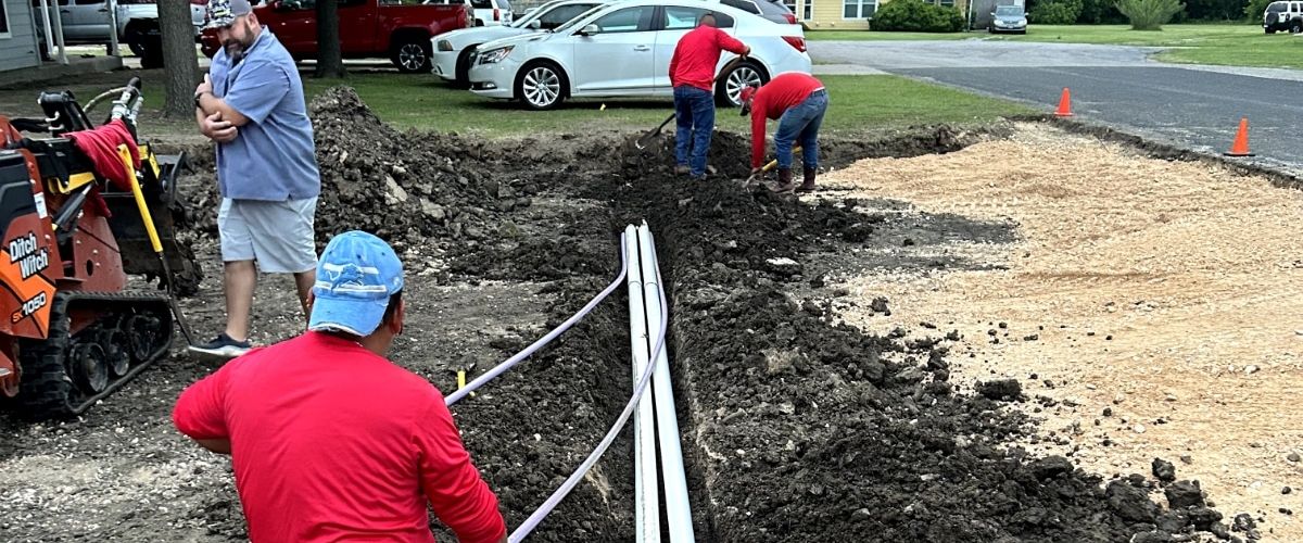 Men working on an outdoor construction project, installing pipes in a trench with a compact utility loader and various vehicles in the background